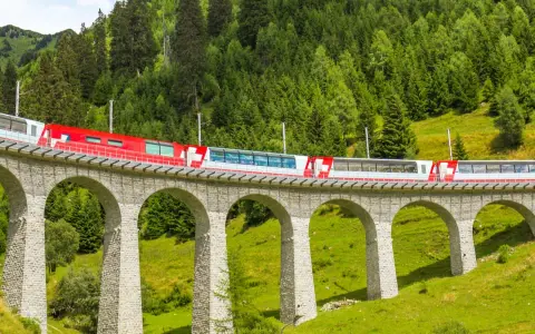 A red and white train travels across a stone viaduct, winding through a lush green valley with a dense forest on the mountainside. This scenic route is part of the Amsterdam to Basel train journey.