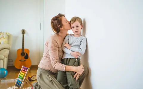 Madre besa a su niño independiente, apoyado en la pared. Juguetes y guitarra en el fondo.