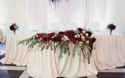 Elegancki bordowy wystrój sali weselnej z bogatą dekoracją kwiatową na stole.