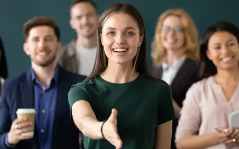 Uśmiechnięta kobieta wyciąga rękę na powitanie. To jej pierwszy dzień w nowej pracy, a zespół wygląda na przyjazny.