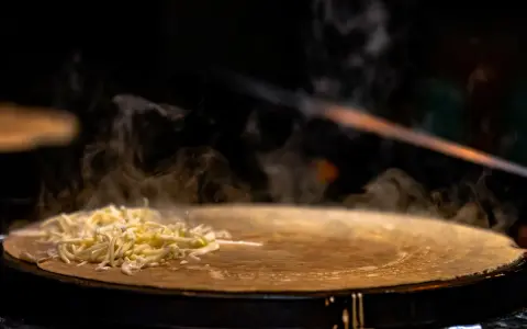 Fromage râpé fondant sur une crêpe chaude, vapeur s'élevant. Une tâche de nettoyage crêpière Tefal brûlée est visible sur le bord.