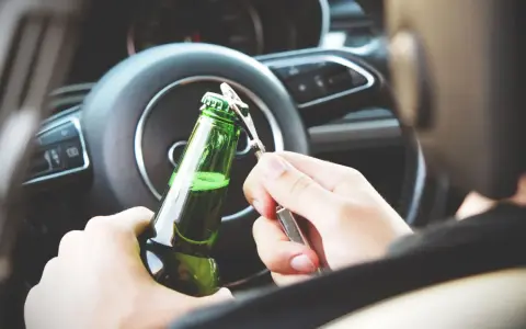 Ręce otwierające butelkę piwa za kierownicą. Pamiętaj, że od 0,2 promila zabierają auto.