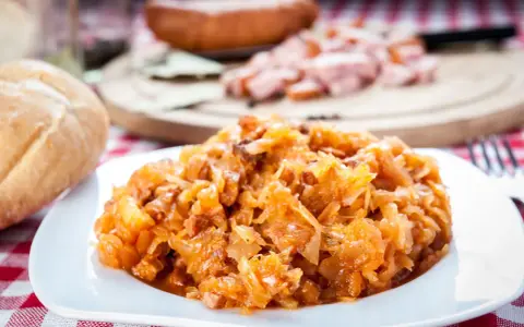 Pyszny bigos Ani, gotowany z pasją, na talerzu. Obok bułka i kawałki kiełbasy.