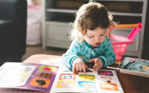 Mała dziewczynka z niepełnosprawnością intelektualną z zaangażowaniem przegląda książeczkę z obrazkami, wskazując paluszkiem na kolorowe ilustracje.