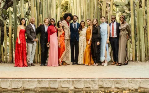 A diverse group of people pose in front of tall cacti, showcasing a vibrant summer color palette with dresses in red, pink, orange, and gold, alongside suits in blues and browns.
