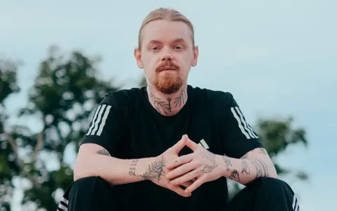 Mężczyzna z rudą brodą i blond włosami, ubrany w czarną koszulkę z trzema paskami. Na szyi i rękach widoczne tatuaże, w tym klucz i napis "kuqe 2115".