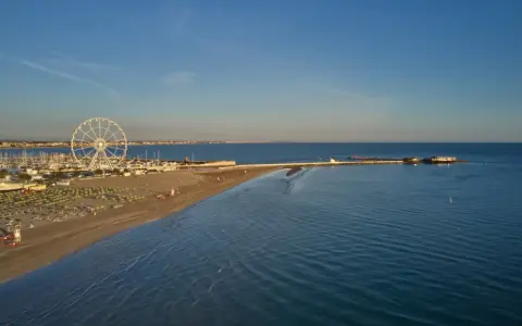 Vista aerea di una spiaggia di Rimini con ruota panoramica, porto turistico e mare calmo sotto un cielo azzurro.