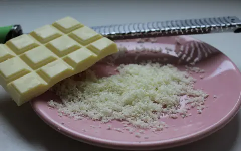 Une râpe à fromage argentée est posée sur une assiette rose, recouverte de copeaux de chocolat blanc fraîchement râpé.