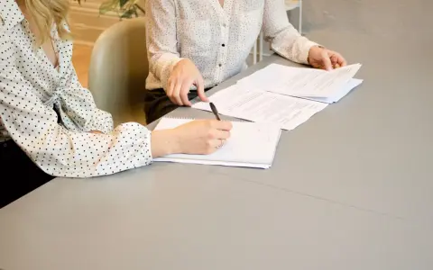 Dwie kobiety omawiają umowę o świadczenie usług. Jedna pisze notatki, druga przegląda dokumenty.
