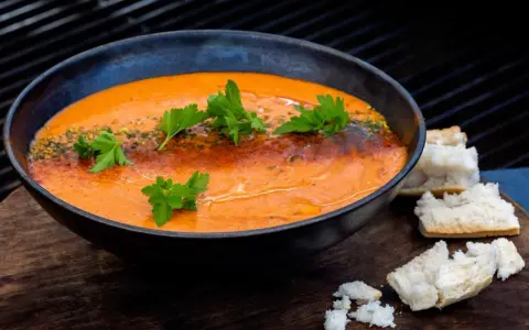En skål med krämig, orange soppa, troligen en smakrik mojo rojo, garnerad med persilja och serverad med brödbitar.
