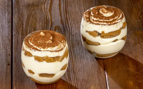 Due bicchieri di tiramisù senza uova, stratificati con crema e savoiardi, spolverati di cacao su un tavolo di legno.