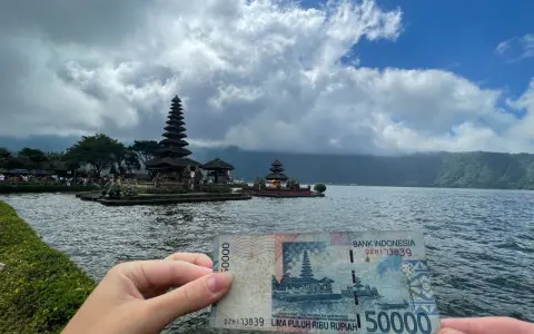 Ręka trzyma banknot 50 000 rupii na tle świątyni na wodzie. Czy Indonezja jest bezpieczna? Tak, podróżowanie po niej jest bezpieczne.