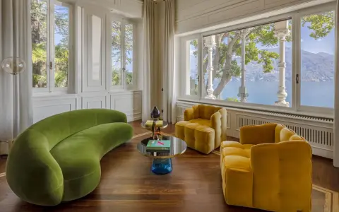 Salotto con divano verde curvo e poltrone gialle, tavolini di design e vista lago. Un'elegante casa Art Déco.