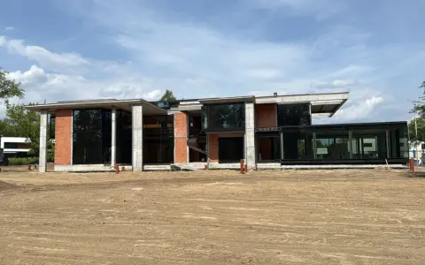 Nowoczesny dom w stanie surowym zamkniętym, z dużymi przeszkleniami i ceglanymi elementami, otoczony placem budowy.