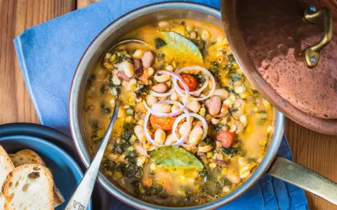 Zuppa ricca con fagioli, verdure e spezie, servita in una pentola di rame. Un piatto confortante di acqua cotta, guarnito con cipolla rossa e alloro.