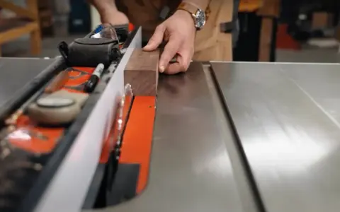Hands guide a wooden plank along a table saw's fence, demonstrating how to use a table saw for precise cuts.