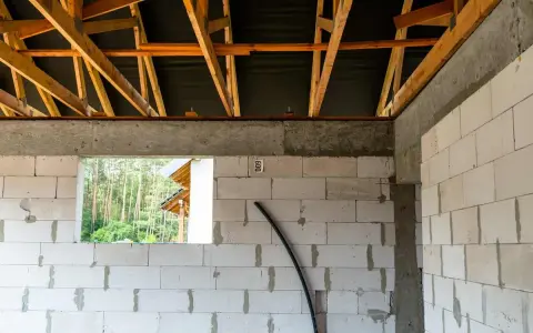 Budowa domu w toku. Widoczny wieniec stropowy, ściany z bloczków, drewniana konstrukcja dachu i otwór okienny z widokiem na las.