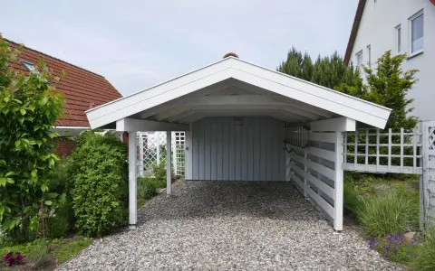 A white wooden carport with a gravel driveway, surrounded by greenery and a white lattice fence. This image shows a finished carport, a great example of how to build a carport.