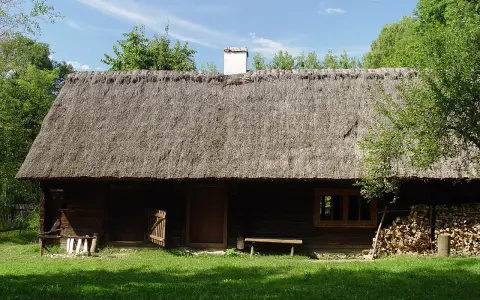 Drewniana chata z krytym strzechą dachem, część ekspozycji Muzeum Wsi Opolskiej. Obok stos drewna i ławka.