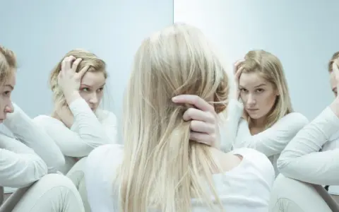 Blondynka w białej bluzie, zmagająca się z kwetaplex, widzi swoje odbicia w lustrze, każde z innym wyrazem twarzy.