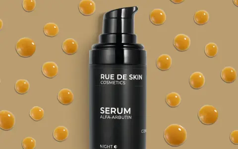 Serum z alfa-arbutyną od RUE DE SKIN. Krople serum unoszą się na beżowym tle, tworząc efekt kropli rosy.