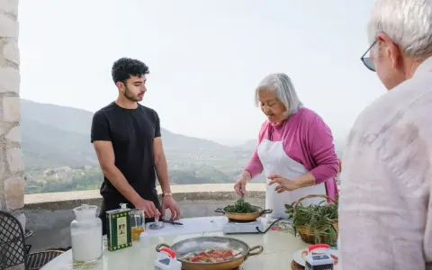 An older woman in an apron cooks greens while a young man and an older man stand nearby on a terrace with a scenic view.