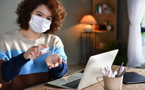 Kobieta w masce dezynfekuje dłonie płynem ze sprayu przy laptopie.
