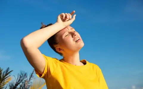 Une jeune femme en t-shirt jaune, le visage marqué par la chaleur, essuie son front sous un ciel bleu intense. La durée d'une insolation est palpable.