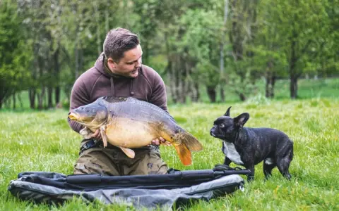 Ein Angler präsentiert stolz einen großen Karpfen, während sein kleiner Hund neugierig zusieht. Ein erfolgreicher Tag beim feedern auf Karpfen.