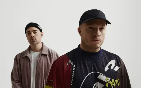 Dwóch mężczyzn, jeden w czapce beanie, drugi w czapce z daszkiem. Na koszulce jednego z nich widoczny jest **guzior fala tekst**.