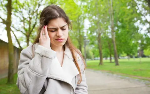 Kobieta w parku trzyma się za głowę, odczuwając ból, jakby strzelanie w uchu przy przełykaniu.