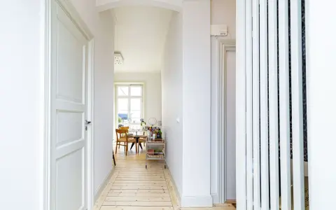 Ljus hall med trägolv och vita dörrar, perfekt för en renovera hall. Genom en valvöppning syns ett runt bord med stolar och ett fönster.