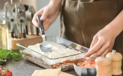 Osoba nakłada sos na warstwy makaronu i mięsa, przygotowując do pieca lasagne.