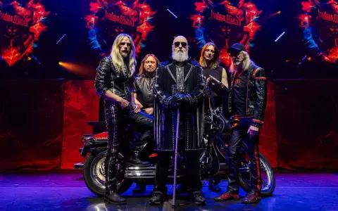 Medlemmar av Judas Priest poserar på scenen med en motorcykel. Bandets logotyp syns i bakgrunden.