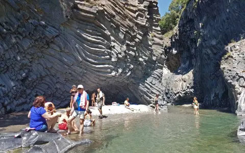 Persone si rilassano e si rinfrescano nelle acque cristalline delle cascate dell'Alcantara, circondate da imponenti pareti rocciose basaltiche.