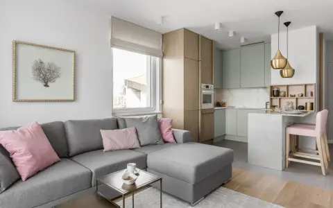 Nowoczesny salon z aneksem kuchennym: szara sofa, pastelowe poduszki, drewniane meble i marmurowa wyspa. Idealne jak urządzić mały salon z aneksem.