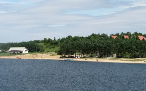 Plaża nad jeziorem Chańcza tętni życiem. Ludzie odpoczywają na piasku, kąpią się w wodzie, a w tle widać domki i las.