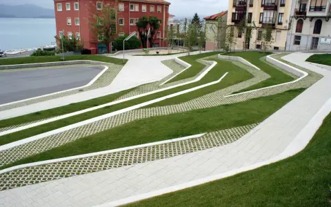 Diseño moderno con césped y caminos de hormigón que forman patrones ondulados. El suelo drenante permite el paso del agua, integrándose con el entorno urbano y el mar al fondo.