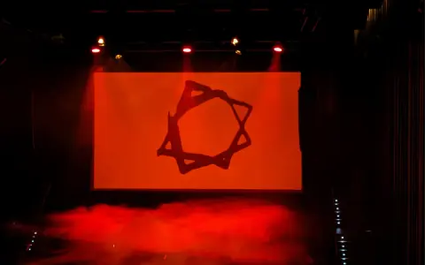 Símbolo estilizado en pantalla naranja, con humo rojo y luces de escenario. Podría ser parte de una dellafuente biografia visual.