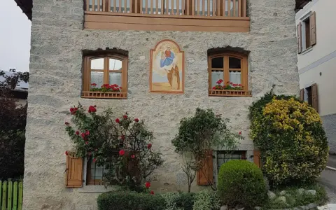 Un affresco sacro adorna un muro di pietra di un borgo trentino alto adige, con rose rosse che sbocciano rigogliose.