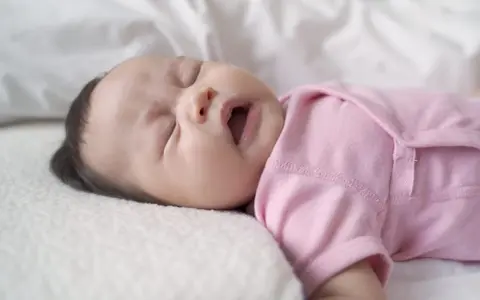 Ein süßes neugeborenes Baby in rosa Strampler niest. Die Augen sind geschlossen, der Mund leicht geöffnet.