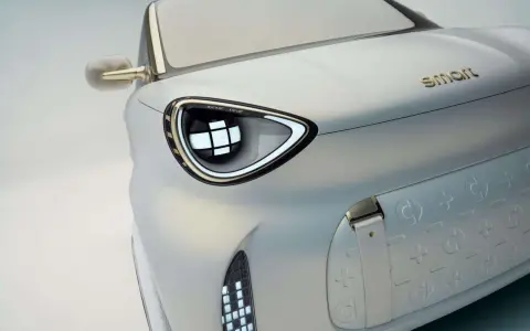 Dettaglio di un'auto elettrica Smart, con un faro futuristico e il logo dorato. Nuovi modelli in arrivo.