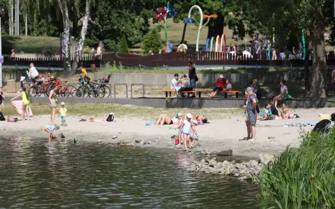 Rodzina odpoczywa nad wodą na piaszczystej plaży Jeziora Paprocany. Dzieci bawią się w piasku i przy brzegu, dorośli relaksują się na kocach i ławkach.