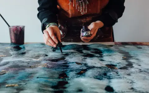 Une artiste crée une **aquarelle sur toile** aux teintes bleues et dorées, ajoutant des touches d'encre avec un compte-gouttes.