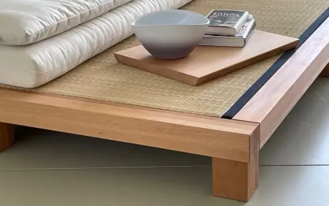 Eine gestapelte, weiße Matratze eines japanischen Bettes ruht auf einer Tatami-Matte. Daneben liegen ein Buch und eine Schale auf einem Holzbrett.