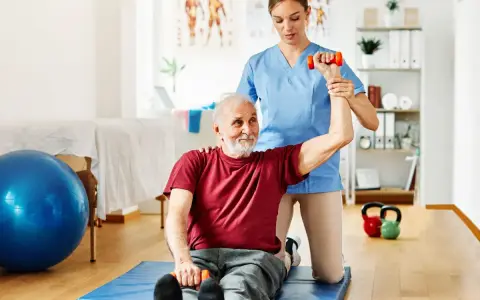 Starszy pan ćwiczy z fizjoterapeutką, podnosząc hantle. To przykład, dla kogo przeznaczony jest bon senioralny – na rehabilitację i aktywność.
