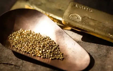 Piccoli granelli scintillanti di oro riciclato riposano in una paletta di rame, accanto a lingotti dorati.