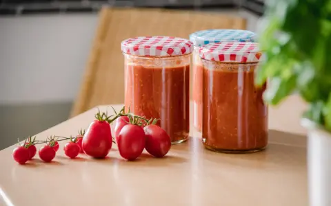 Frische Tomaten und eingekochte Tomatensauce in Gläsern. Die Frage "Tomatensauce einkochen wie lange?" beschäftigt viele Hobbyköche.