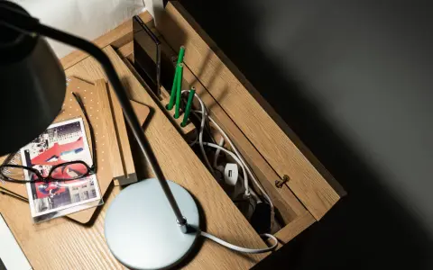 Drewniana szafka nocna z przegródkami, gdzie można schować wystające kable ze ściany. Na blacie lampka, okulary i notatnik.