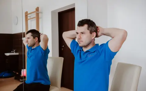Mężczyzna w niebieskiej koszulce wykonuje ćwiczenia na kręgosłup szyjny, opierając dłonie na karku i delikatnie napinając mięśnie.
